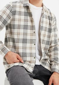 Camicia button-up con motivo a quadri beige e neri, realizzata in tessuto morbido, con colletto a punta e maniche lunghe. Indossata sopra una camicia bianca.