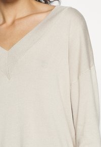 Pull tricot beige avec col en V et larges manches, présentant une texture côtelée fine et un design minimaliste.