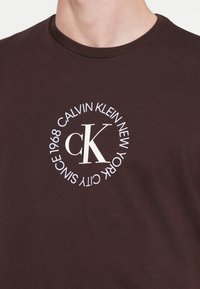 Pruun puuvillane t-särk, millel on valge ringikujuline logo, kus on kiri "CALVIN KLEIN NEW YORK" ja "SINCE 1968," keskel stiliseeritud "cK".