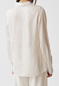 Långärmad, ljuskrämsfärgad skjorta med en slät yta, knäppning vid ärmsluten och klassisk krage. Har en något rundad nederkant.