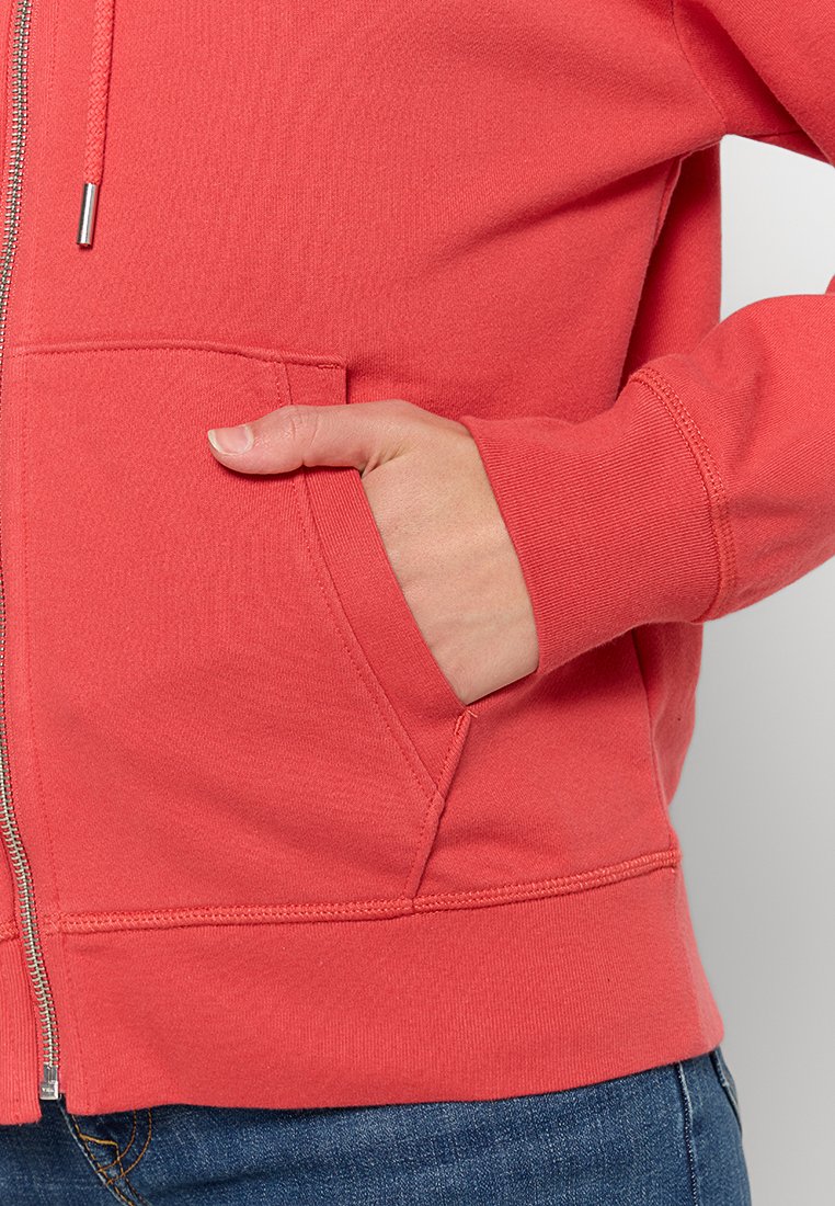 Felpa con zip corallo realizzata in tessuto testurizzato, dotata di tasca frontale e polsini a coste. Dettagli visibili della zip e delle cuciture.