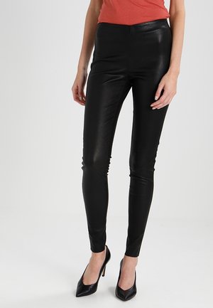 Femme portant un pantalon moulant en similicuir noir et des talons hauts noirs, debout devant un fond clair uni.
