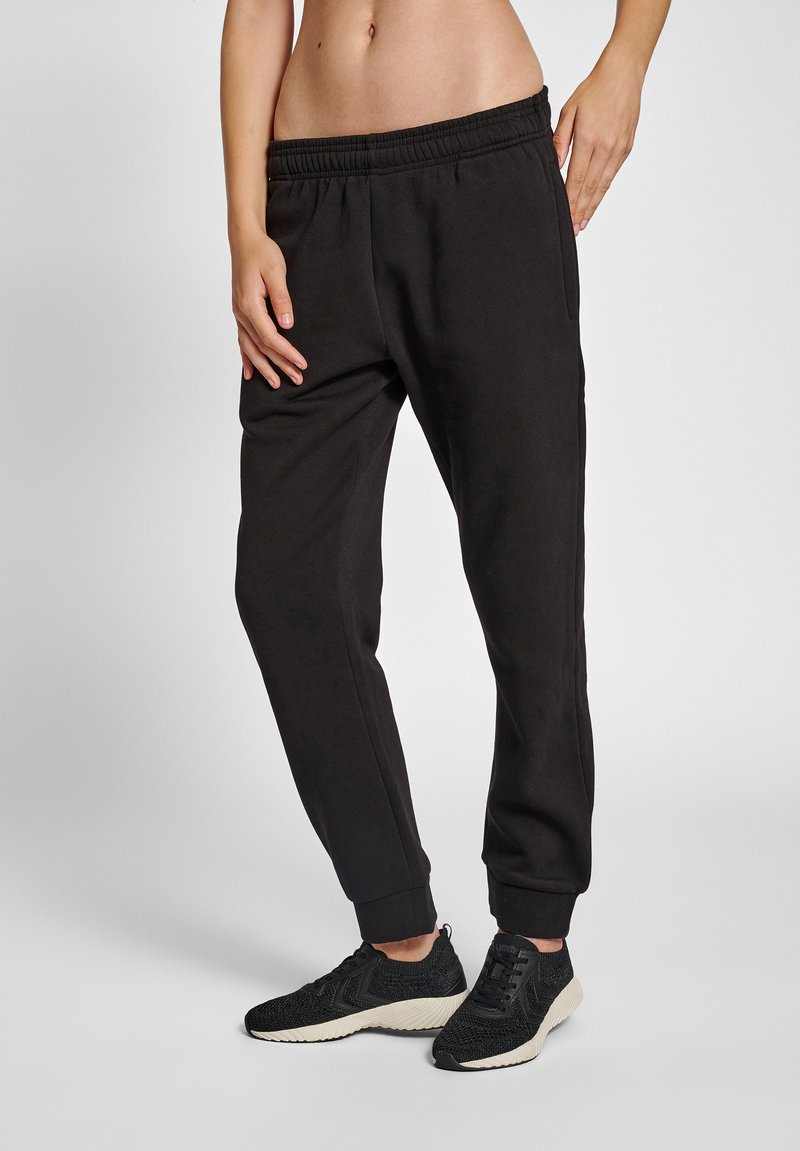 Pantalones de chándal negros con cintura y puños elásticos; tejido suave y liso; combinados con zapatillas deportivas negras de textura.