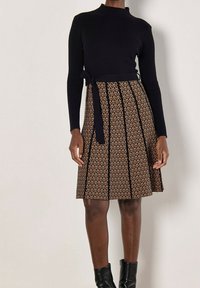 Femme portant un haut noir côtelé à manches longues associé à une jupe mi-longue à motifs marron et noirs et des bottines noires.