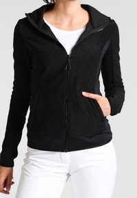 Zwarte zip-up jas met een ruitvormig quiltpatroon en zijzakken. Heeft een capuchon en een aansluitend design. G gedragen over een witte top.