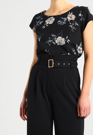 Žena oblečená v černé květované krátkosměrné blůze vsazené do vysokých černých kalhot s širokým opaskem a zlatou sponou.