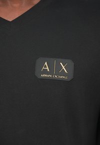 Μαύρο T-shirt με V λαιμόκοψη, φτιαγμένο από μαλακό ύφασμα, με καρέ λογότυπο που έχει το "A|X" και "Armani Exchange" σε χρυσά γράμματα.