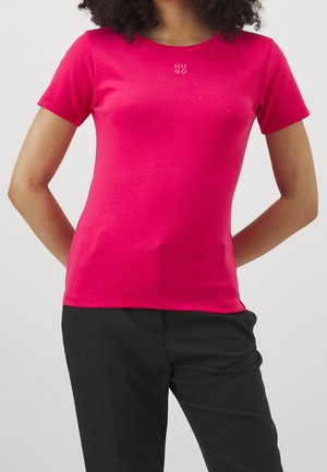 Kvinde iført lyserød tætsiddende T-shirt med korte ærmer og lille "HUGO" logo på brystet samt sorte bukser, hænderne bag ryggen.