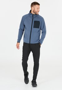 Grijze zip-up jas met een zwarte borstzak, gecombineerd met zwarte tapered pants en zwarte schoenen. Soepele stof met een getailleerd ontwerp.