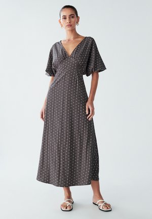 Ilga suknelė tamsiai rudos spalvos audinyje su baltais taškeliais, su V formos iškirpte, laisvomis rankovėmis ir laisva silueto forma. Paprastos plokščios sandalės.