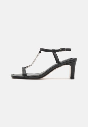 Black leather heeled sandals featuring a T-strap design, metal accents, a slender heel, and a smooth finish with an open toe.