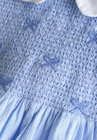 Megzta mėlyna suknelė su raukšlėtu liemeniu, pasižyminti tekstūruotu audimu. Apačioje yra vertikalios mėlynos ir baltos juostos. Papuošta dekoratyvia siuvinėjimu.