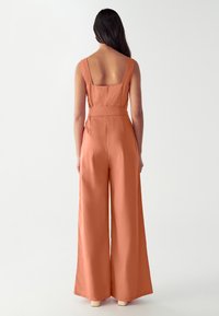 Žena s dlouhými tmavými vlasy, oblečená v bezramínkovém, čtvercovém, širokém kombinézovém overalu v měděné barvě, stojící tak, že je otočená zády k pozorovateli.