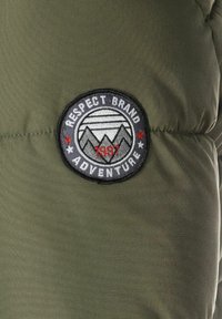 Grøn nylonjakke med et syet rundt mærke. Mærket viser bjerge, teksten "RESPECT BRAND ADVENTURE" og året "1997."