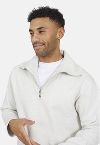 Lichtgrijze ritspull met een zachte stof, voorzien van een hoge kraag, een rits van kwartlengte en ribbelaccenten langs de opening.