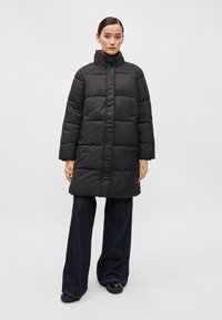 Črn napihnjen plašč z visokim ovratnikom, ki ima kvadratni vzorec, dolge rokave, stranske žepke in ravno krojno do dolžine sredine stegen.