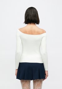 Bela rebrasta majica z dolgimi rokavi, spuščena z ramen, v kombinaciji z mornarsko modro krilo s pliseji in vzorčastimi nogavicami, prikazana od zadaj.