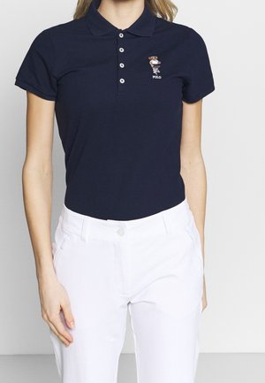 Kobieta w granatowej koszulce polo z logo niedźwiedzia i białych spodniach, stojąca na jednolitym jasnym tle.