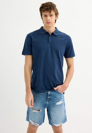 Camiseta polo azul marino con cuello y mangas cortas, combinada con pantalones cortos de mezclilla desgastados, con dobladillos sin terminar y rasgaduras visibles.
