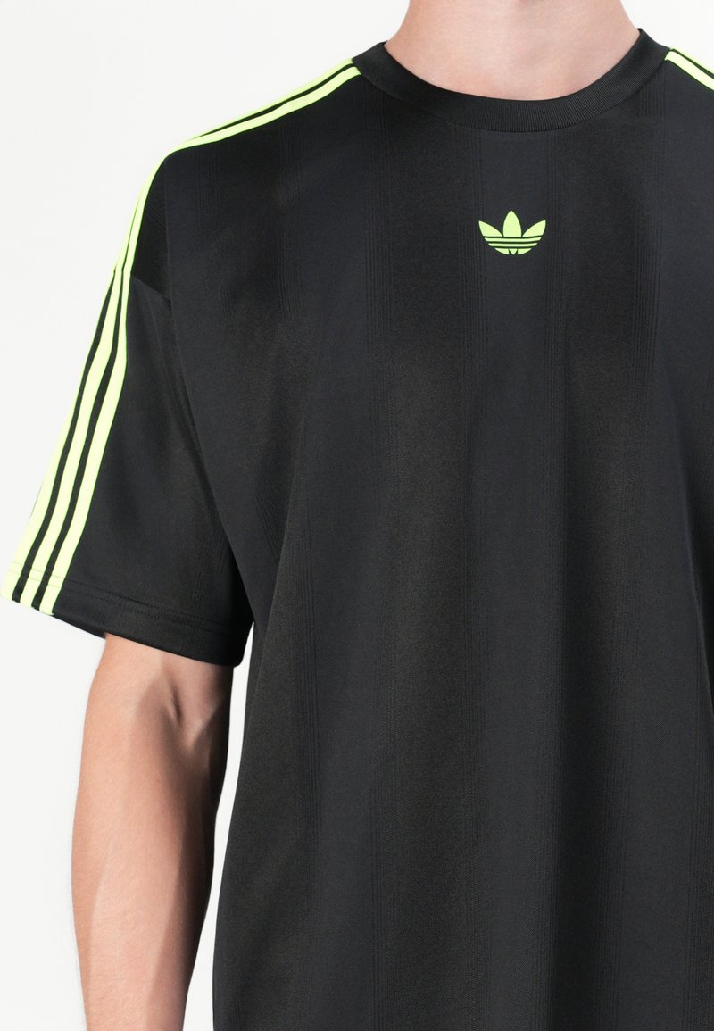 Camisa negra de manga corta con cuello redondo, que presenta tres rayas verticales en amarillo brillante en los hombros y un pequeño logo en el pecho.