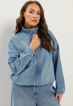 Femme aux longs cheveux bruns portant une veste en jean bleue ample et un jean, tenant le col de la veste d'une main.