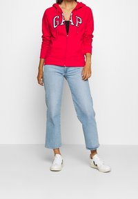 Sudadera roja con cremallera y logo de "GAP", combinada con jeans cortos de mezclilla azul claro y zapatillas blancas. La sudadera tiene un cordón y una textura suave.