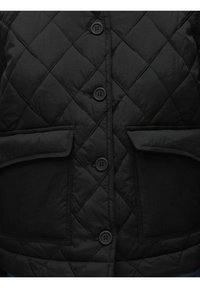 Juoda siūlų pūkinė striukė su mygtukų uždarymu, dviem didelėmis priekinėmis kišenėmis ir glotnia, šiek tiek blizgia tekstūra.