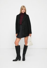 Crni vuneni kaput sa krojem po mjeri, burgundski turtleneck, crna plisirana suknja i crne kožne čizme do koljena. Bijela torba s kratkim ručkama.