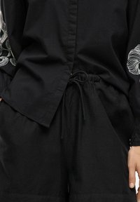 Camicia nera in cotone con dettagli ricamati sulle maniche, abbinata a pantaloni corti neri con coulisse. Presenta una vestibilità rilassata e una texture morbida.