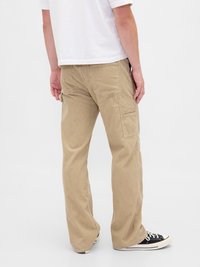 Μπεζ παντελόνι cargo από βαμβακερό μείγμα, με πλαϊνές τσέπες, ευθεία γραμμή και εμφανείς ραφές. Φορεμένο με μαύρα αθλητικά παπούτσια.