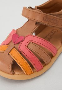 Tan färgad lädersandal med rosa och orange detaljer, med utskärningar och ett hjärtdetalj. Justerbar rem för en säker passform. Mjuk sula.
