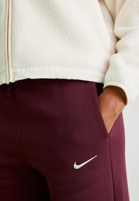 Krämfärgad fleece zip-jacka med en slät krage, i kombination med mörk burgundy sweatpants med en liten vit Nike-logotyp på låret.