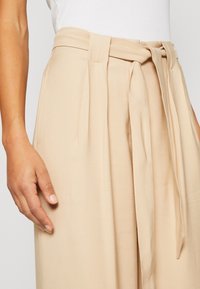 Beige broek met een hoge tailleband, stoffen riem en plooien. Gladde textuur met subtiele glans, getoond met een hand aan de zijkant geplaatst.