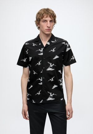 Jeune homme portant une chemise noire à manches courtes avec des motifs d'oiseaux blancs, debout devant un fond blanc uni.