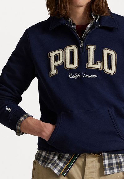 Temno modra majica s kapuco z vezenim napisom "PO LO" in "Ralph Lauren". Ima logo polo na rokavu in dizajn z visokim ovratnikom.