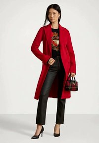 Moteris dėvi raudoną ilgą paltą, juodą grafišką megztinį, juodas kelnes, juodus aukštakulnius bato, laiko mažą languotą rankinę, stovinti prieš paprastą foną.