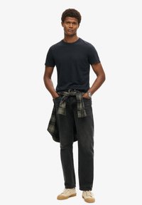 T-shirt nera a maniche corte, jeans in denim scuro, camicia a quadri legata in vita, sneakers color sabbia, posa in piedi, sfondo neutro.