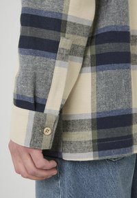 Chemise à manches longues à carreaux présentant des motifs beiges, navy, bleus et verts, fabriquée en coton texturé avec un poignet boutonné.