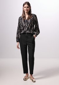 Schwarz gemusterte Bluse mit langen Ärmeln, kombiniert mit taillierten schwarzen Hosen und metallischen flachen Schuhen. Einfaches und modernes Design mit subtilen Texturen.