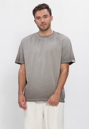 Uomo con capelli castani corti che indossa una maglietta grigia larga e pantaloni bianchi, in piedi su uno sfondo bianco uniforme.