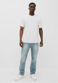 Camiseta blanca de cuello redondo, jeans de color azul claro y zapatillas blancas. El modelo está de pie recto frente a un fondo neutro. Diseño simple, ajuste informal.
