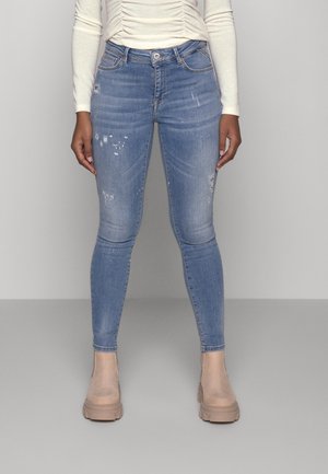 Personne portant un jean skinny bleu clair usé, des bottines beiges et un top blanc à manches longues, debout devant un fond uni.