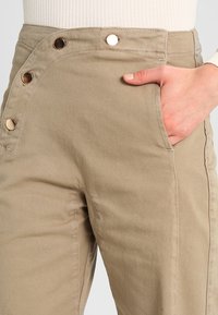 Acercamiento a unos pantalones beiges con botones a presión en el lateral y una mano descansando en el bolsillo delantero, combinados con una blusa blanca de canalé.