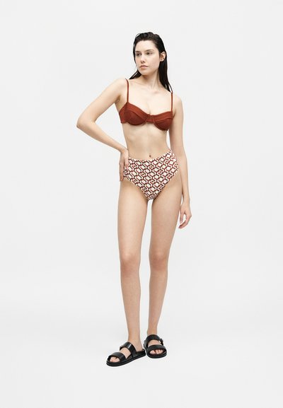 Γυναίκα που στέκεται, φοράει τοπ μαγιό σε σκουριασμένο χρώμα, σλιπ μαγιό με υψηλή μέση και μαύρα σανδάλια σε λευκό φόντο.