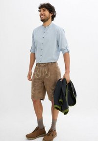 Uomo con camicia azzurro chiaro a bottoni con maniche arrotolate, pantaloni corti in pelle marrone ricamati, scarpe marroni, che tiene un maglione scuro con rifiniture verdi.