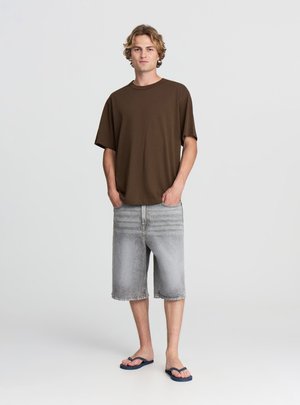 Giovane uomo in piedi con le mani in tasca, indossa una maglietta marrone, pantaloncini di jeans grigi fino al ginocchio e infradito blu navy.