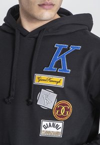 Felpa nera con cappuccio presenta toppe ricamate: 'K' blu, 'Gianni Kavanagh' giallo, 'K' grigio ed emblema tondo bordeaux. Tessuto morbido, cordini al cappuccio.