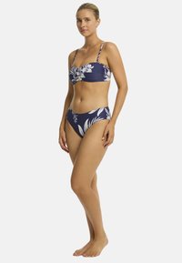 Kobieta w kwiecistym bikini stoi pewnie na białym tle, prezentując wzór i kolory kostiumu kąpielowego.