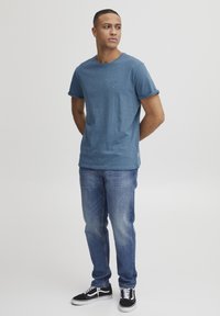 Blå t-skjorte med korte ermer og rullede ermer, med en liten logo på brystet. Matchet med lyseblå jeans og svarte joggesko.