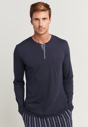 Mann mit hellbraunen Haaren, der ein langärmeliges navyfarbenes Henley-Shirt mit zwei Knöpfen und einer Brusttasche vor einem schlichten Hintergrund trägt.